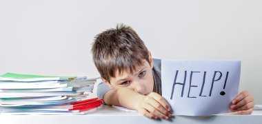Sad tired frustrated kid sitting at the table with many books and holding paper with word Help. Learning difficulties, education, online learning, school at home