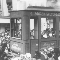 Die Anfänge des Brezelumzugs – ein „Eilwagen Speyer-Dudenhofen“ mit festlich gekleideten Insassen, gezogen von Pferden, rollt durch die Menge der Schaulistigen in der Maximilianstraße, 1911.