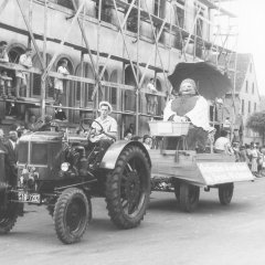 Noch einmal ein Blick in die Gilgenstraße: Ein Traktor zieht einen Motivwagen des Festzugs mit einer volkstümlichen „Brezelfrau“ und einem ihr gewidmeten Sinnspruch, 1955.