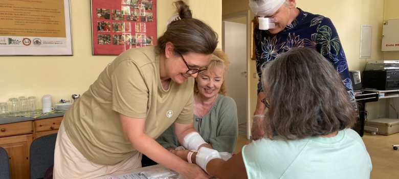 4 Personen auf dem Bild, eine Person sitzt am Tisch, zwei Personen verbinden mit Verbandsmaterial ihren Arm. Eine weitere Person steht daneben. Sie trägt einen Kopfverband und ein Pflaster auf der Nase.