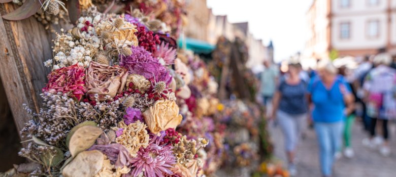 Florale Dekoration an einem verwitterten Holzmast, im Hintergrund geschäftiges Treiben auf der Maximilianstraße in Speyer.