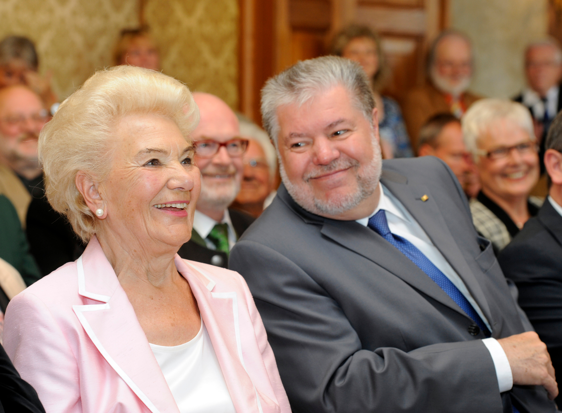 Margarete Boiselle-Vogler sitzt links im Bild in einem hellrosa Blazer und lächelt. Rechts neben ihr sitzt Kurt Beck in einem grauen Anzug. Im Hintergrund sind weitere Gäste zu sehen.