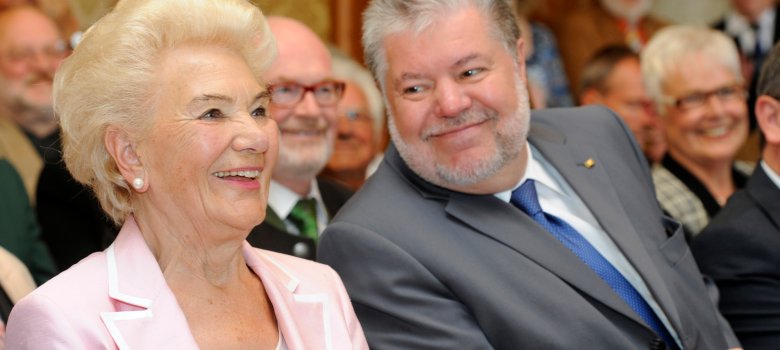 90. Geburtstag Margarete Boiselle-Vogler Speyer Margarete Boiselle-Vogler sitzt links im Bild in einem hellrosa Blazer und lächelt. Rechts neben ihr sitzt Kurt Beck in einem grauen Anzug. Im Hintergrund sind weitere Gäste zu sehen.