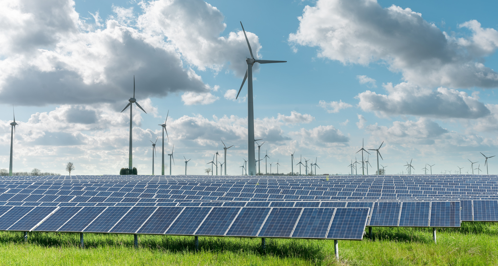 Ein Solarpark mit zahlreichen Solarmodulen auf einer grünen Wiese. Dahinter stehen mehrere Windkraftanlagen. Am Himmel sind große, teils dichte Wolken vor blauem Himmel zu sehen. Das Bild zeigt die Kombination von Solar- und Windenergie zur Erzeugung erneuerbarer Energie.