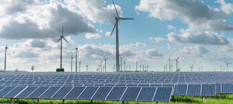 Ein Solarpark mit zahlreichen Solarmodulen auf einer grünen Wiese. Dahinter stehen mehrere Windkraftanlagen. Am Himmel sind große, teils dichte Wolken vor blauem Himmel zu sehen. Das Bild zeigt die Kombination von Solar- und Windenergie zur Erzeugung erneuerbarer Energie.