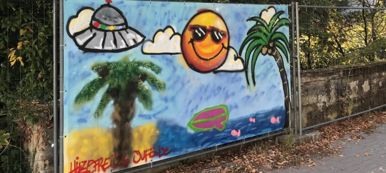 Bauzaunbanner, der durch Jugendliche im Graffiti-Workshop gestaltet wurde. Er zeigt einen blauen Himmel, Palmen, eine lächelnde Sonne mit Sonnenbrille sowie ein Ufo.
