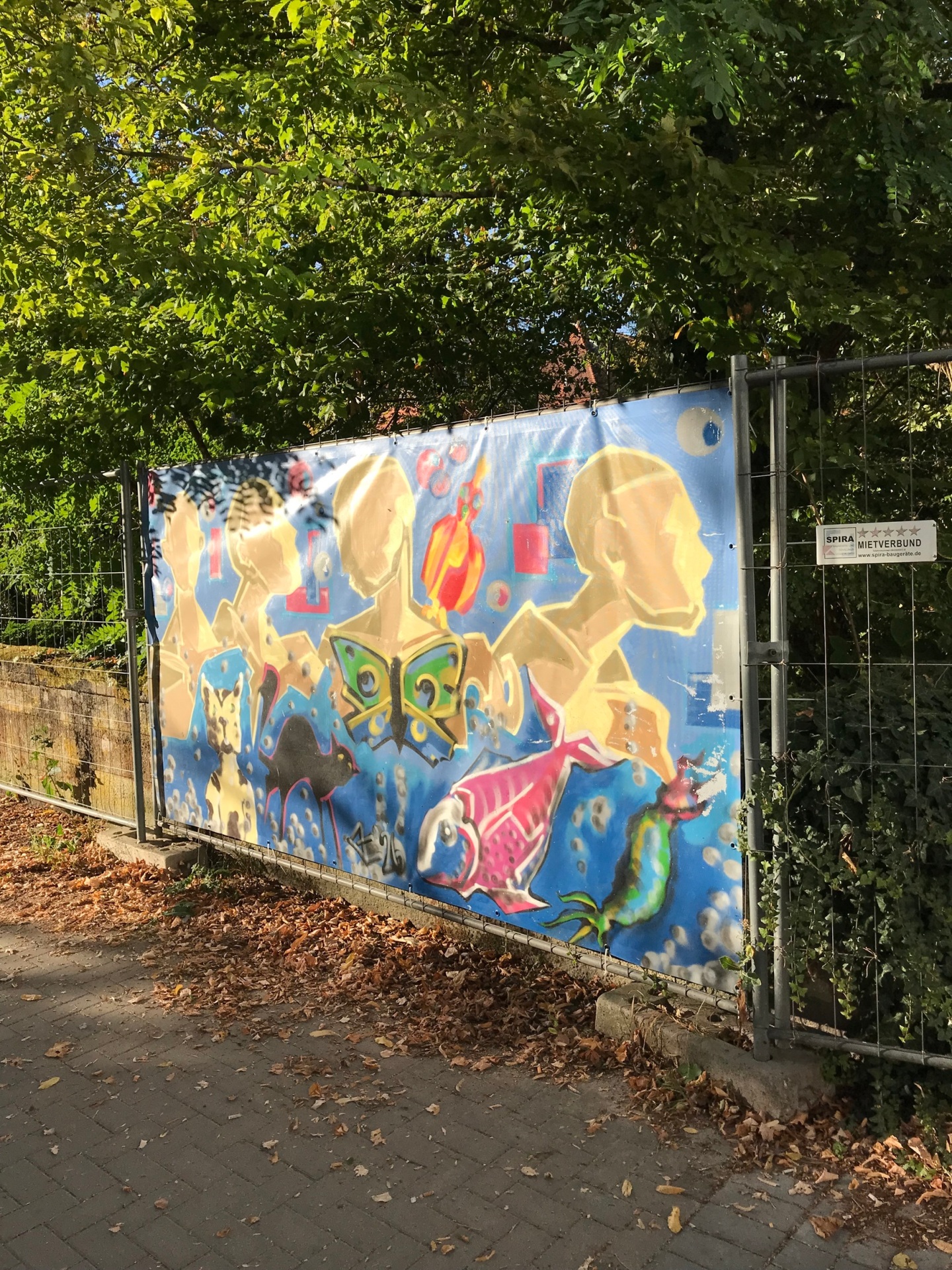 Bauzaunbanner, der durch Jugendliche im Graffiti-Workshop gestaltet wurde. Er zeigt stilisierte Köpfe in Gelb- und Brauntönen sowie einen Koi.
