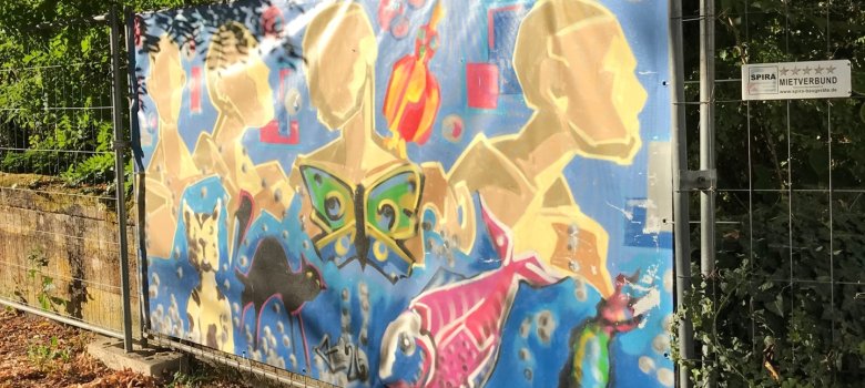 Bauzaunbanner, der durch Jugendliche im Graffiti-Workshop gestaltet wurde. Er zeigt stilisierte Köpfe in Gelb- und Brauntönen sowie einen Koi.