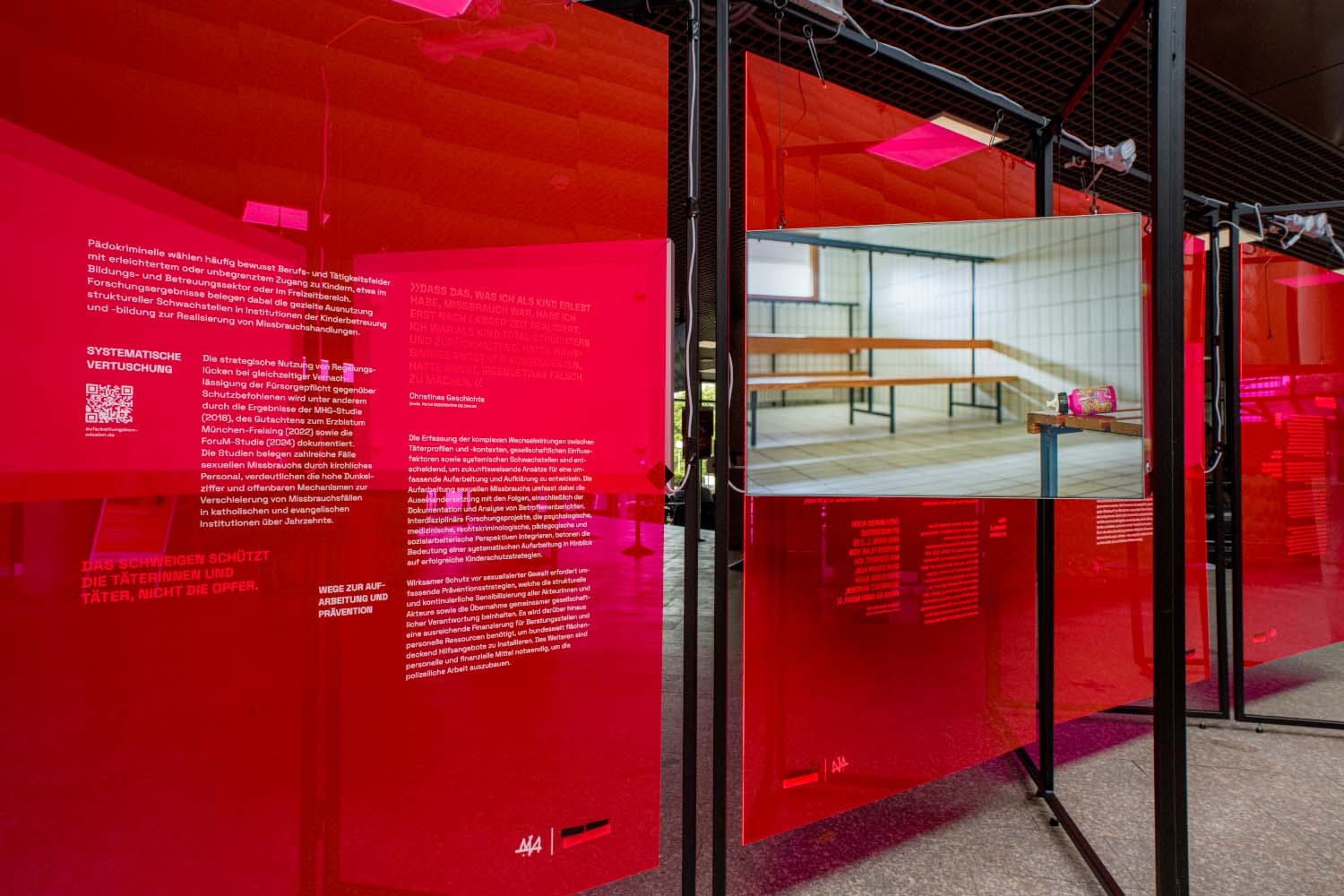 Blick in die Ausstellung „Kunst gegen Missbrauch“ von Nezilla mit transparenten roten Ausstellungstafeln und mehreren Bildschirmen. Die Tafeln enthalten Texte und Fotografien zu sexualisierter Gewalt und ihren Folgen. Im Vordergrund ist auf einem Bildschirm das Innere einer Sporthalle zu sehen.