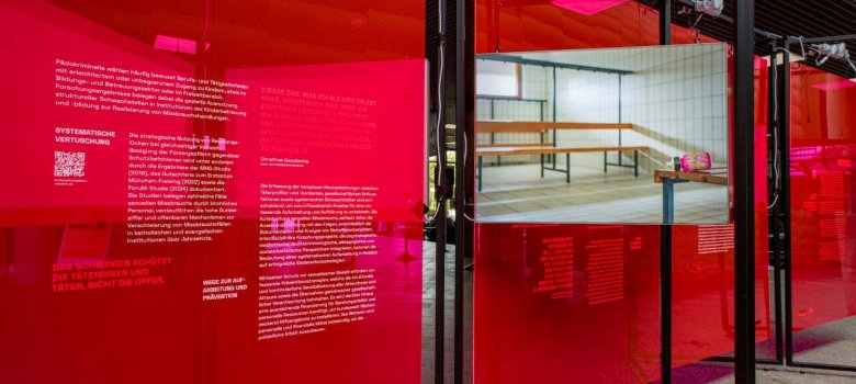 Blick in die Ausstellung „Kunst gegen Missbrauch“ von Nezilla mit transparenten roten Ausstellungstafeln und mehreren Bildschirmen. Die Tafeln enthalten Texte und Fotografien zu sexualisierter Gewalt und ihren Folgen. Im Vordergrund ist auf einem Bildschirm das Innere einer Sporthalle zu sehen.