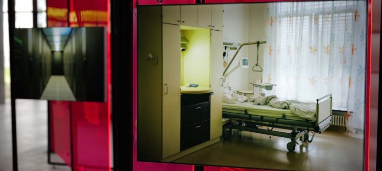 Blick in die Ausstellung „Kunst gegen Missbrauch“ von Nezilla mit roten transparenten Ausstellungselementen. Ein großer Bildschirm zeigt das Foto eines Krankenzimmers mit einem Pflegebett und medizinischer Ausstattung. Im Hintergrund sind weitere Ausstellungstafeln und Bildschirme zu sehen.