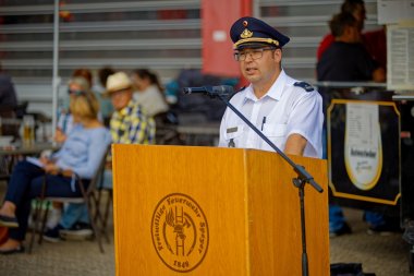 Brand- und Katastrophenschutzinspekteur Peter Eymann steht an einem Rednerpult und spricht zu Menschen. Er trägt die Festuniform der Feuerwehr, ein blauer Hut mit goldfarbener Zierde und ein weißes Hemd. Im Hintergrund hören Personen aufmerksam zu.